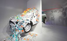 The front half of a car crashing through a wall with white paper (with alphabets) falling off it. The behind of the car is on the other side of the wall with images of people on the wall 