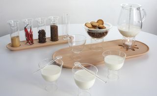 5 Clear cylinder glasses filled with condiments on a beech wood tray. 3 clear glass bowl with milk, with a clear glass cylinder base and a clear stiring spoon hovering about it. A clear glass jug of milk with a thermometer inside it placed on a beech wood tray. A clear glass bowl filled with biscuits with a clear glass cylinder base. Photographed on a white table against a white background