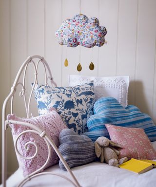 Pastel pink and blue patterned and knitted cushions on a white child's daybed with hanging fabric cloud mobile.