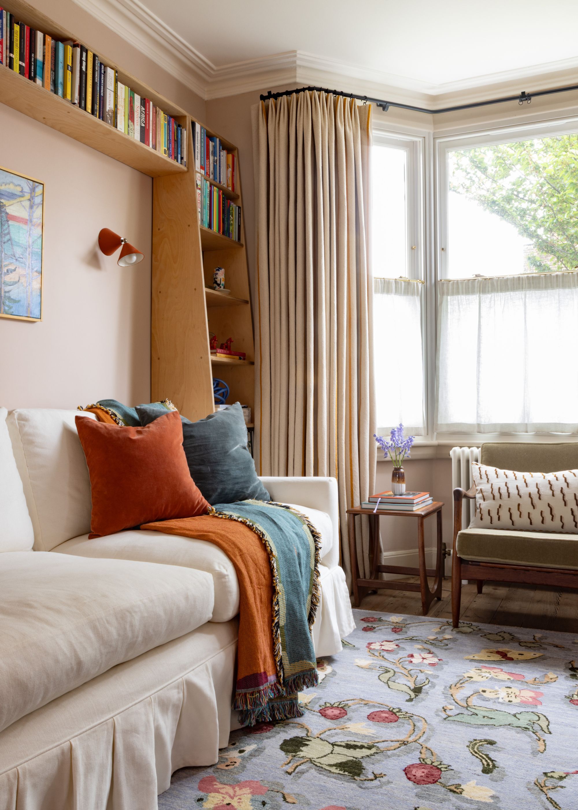 A living room with a bay window, soft peach walls, and a cream sofa. A patterned powder blue large rug, beige curtains, and burnt orange and dark blue throws and cushions on the sofa.