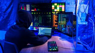 Rear view of computer hacker in dark hall wearing hooded top hacks into a computer network, types software code on a keyboard and monitors a virus attack to hack into network systems and servers.
