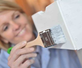 A lady, wearing blue overalls, painting woodwork in the home white, using Osmo