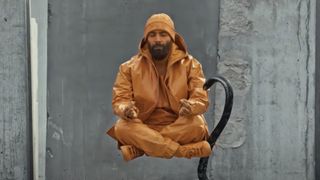 A still from the Netflix TV series A Round of Applause in which a man is dressed in orange and painted orange and he is levitating while in a meditating position with his eyes closed.