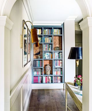 Hallway space with painted cream walls, dark wooden flooring, white painted wooden console table with black and metallic table lamp, looking on to a built in bookshelf painted blue, filled with books and ornaments, two framed pictures on wall with wall lamps beside