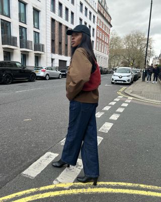 Georgie wears black heeled boots, wide-leg blue jeans, a brown jacket, and red suede shoulder bag.