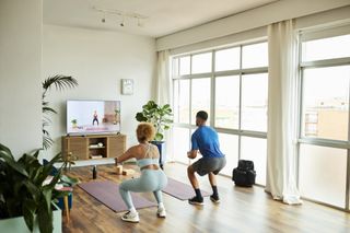 A young couple doing squats in their home while following a workout video on their television.