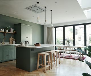 large kitchen diner with green cabinets and walls, white worktops, wooden stools, white dining table with wooden legs and white chairs