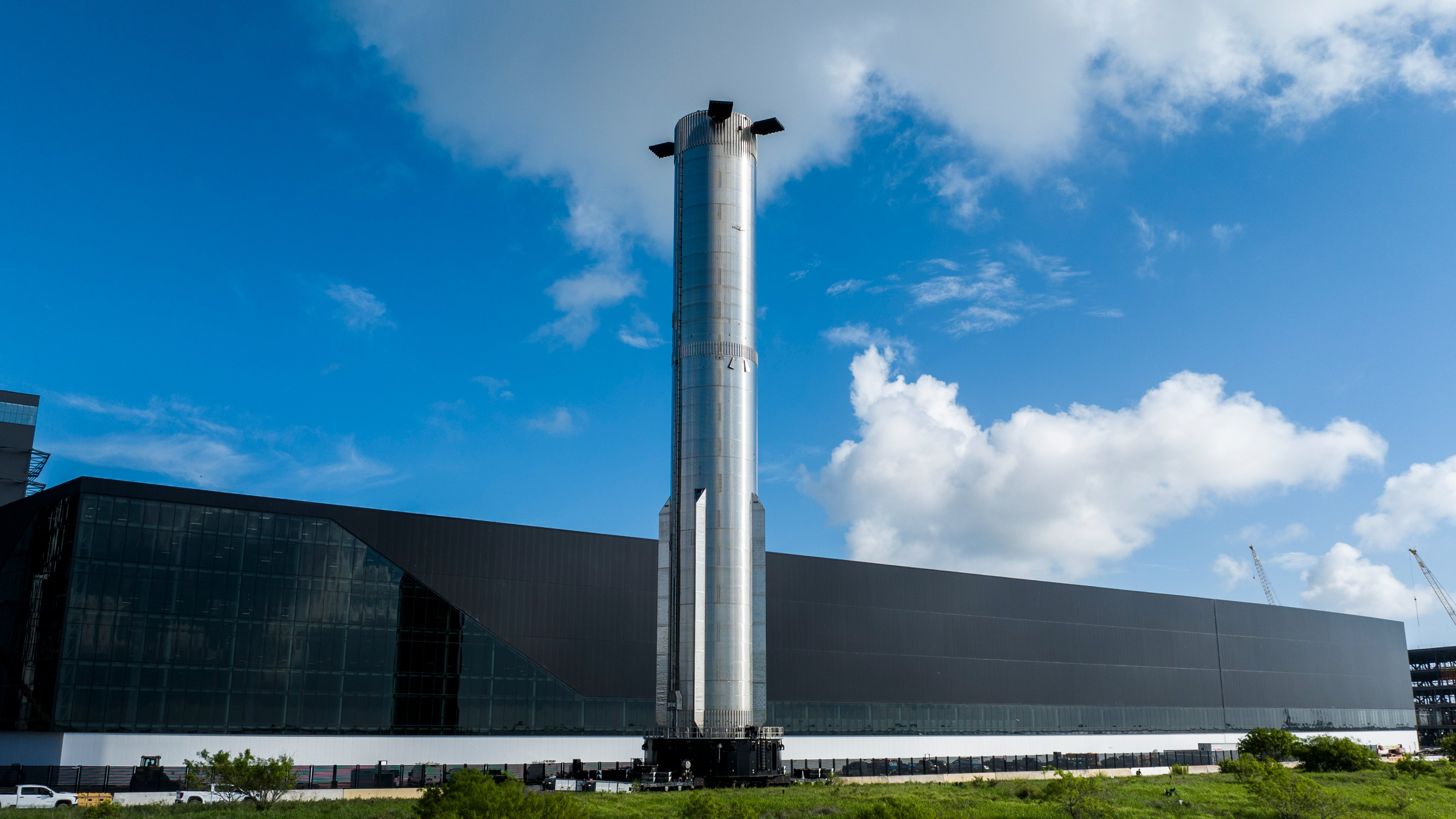 a big silver rocket rolls along a road past a low-slung but very large dark building