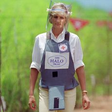 Princess Diana wearing a protective visor and Halo Trust mine vest walking through a field covered with red caution signs