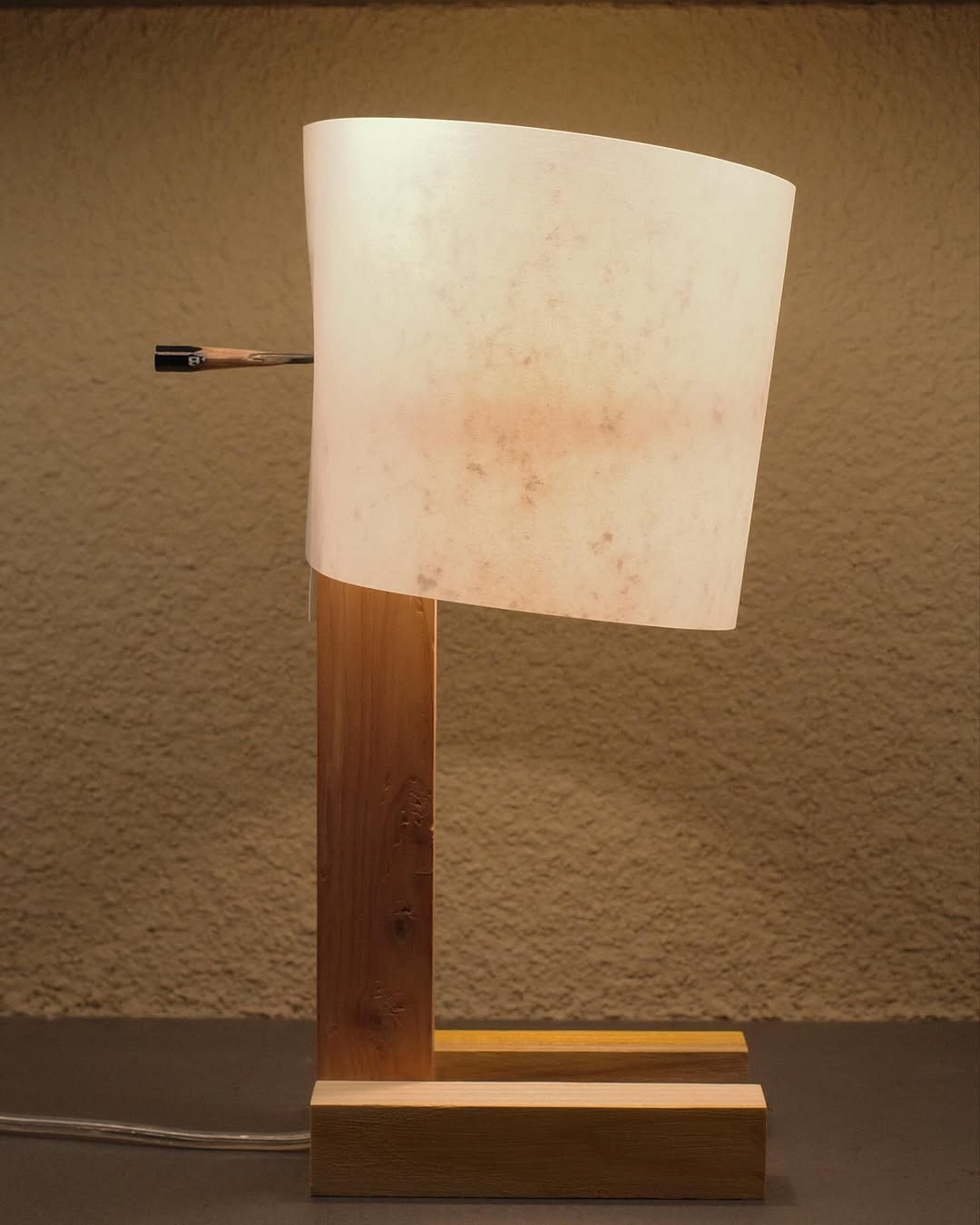 A table lamp with a wooden base and white shade. The shade is held in place by a pencil that has been almost completely sharpened, there is almost only a very spiky tip left
