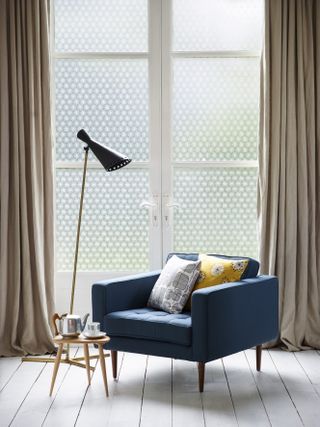 corner of a living room with large open windows covered in a polka dot window film, dressed with neutral coloured curtains and styled with deep blue arm chair and patterned cushions