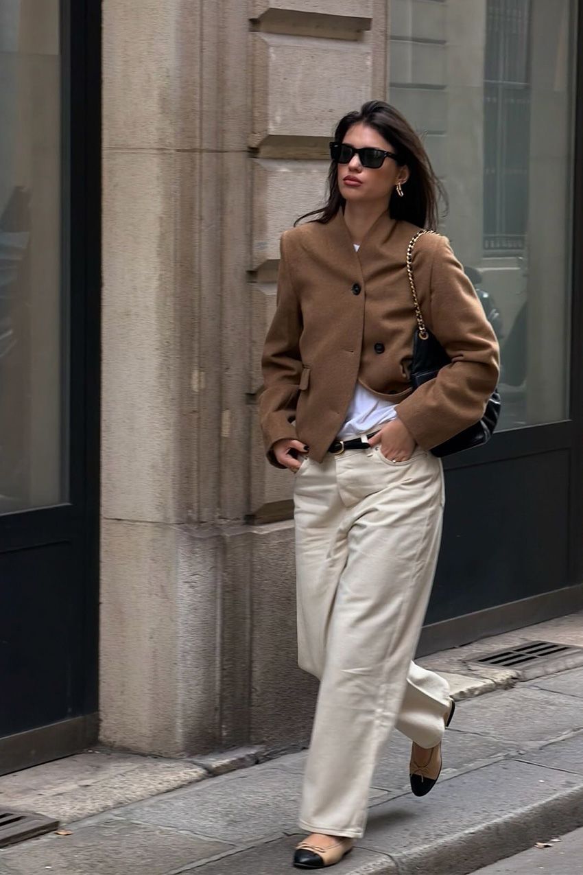 Fall shoe trends in Paris are shown in a photo of a woman on the sidewalk wearing the cap-toe shoe trend styled with a dark brown blazer over a white tee styled with off-white jeans and a black bag
