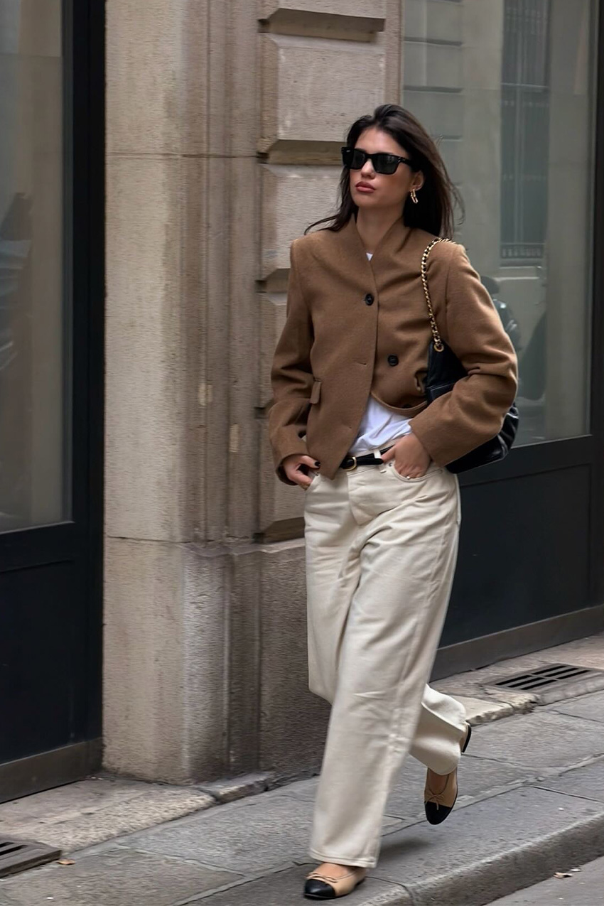 Fall shoe trends in Paris are shown in a photo of a woman on the sidewalk wearing the cap-toe shoe trend styled with a dark brown blazer over a white tee styled with off-white jeans and a black bag