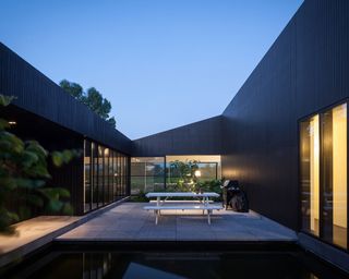 A central courtyard in a residntial home featuring a bbq grill, and white park bench and table on concrete square floor design by a fish pond. surrounded by walls of clad in a dark, heat-treated wood with glass doors