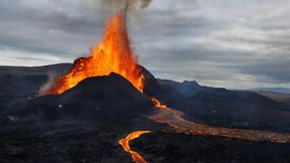 flaming hot magma spews from a crack in the earth, jetting upward in a splash, and streaming down the right of the image over hard, black rock surface.