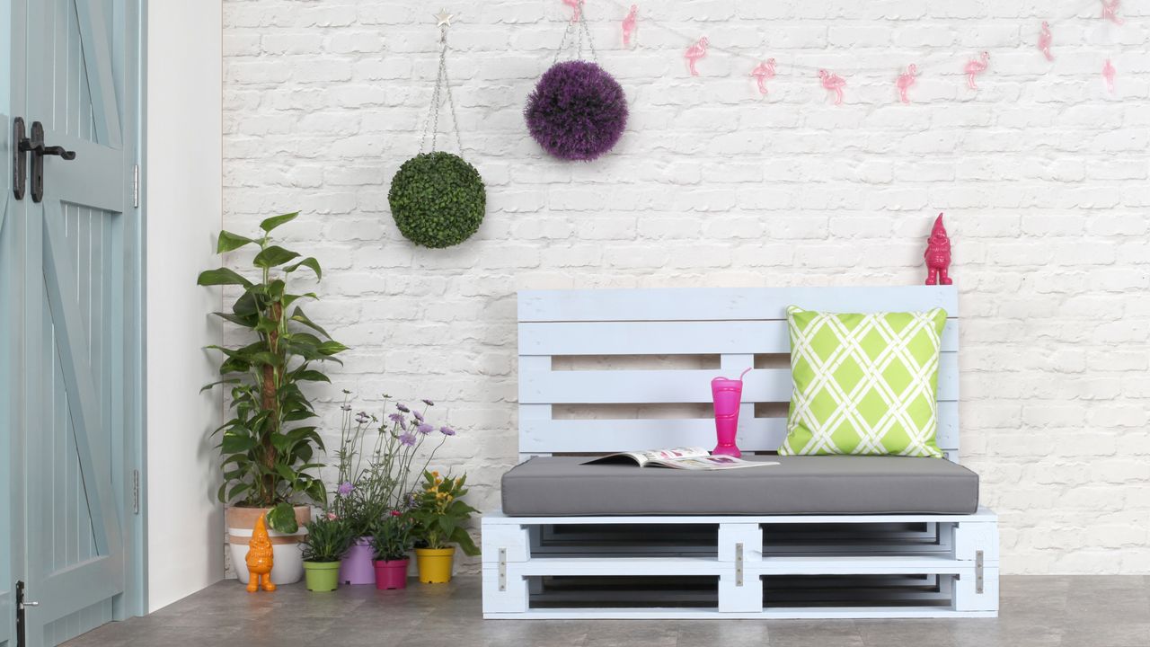 Outdoor pallet furniture in a bench style, painted in a pastel blue with cushions by a wall