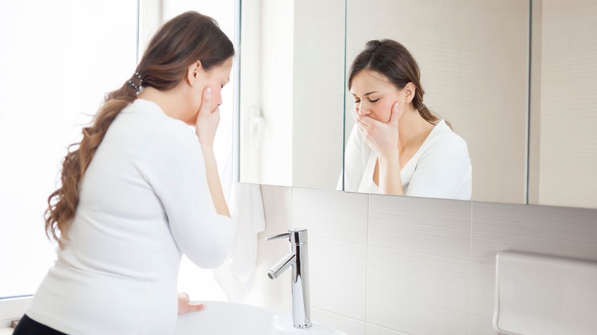 pregnant woman pictured standing in front of a bathroom mirror holding her mouth as if she&#039;s going to be ill