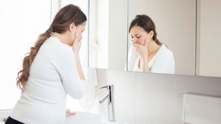 pregnant woman pictured standing in front of a bathroom mirror holding her mouth as if she's going to be ill