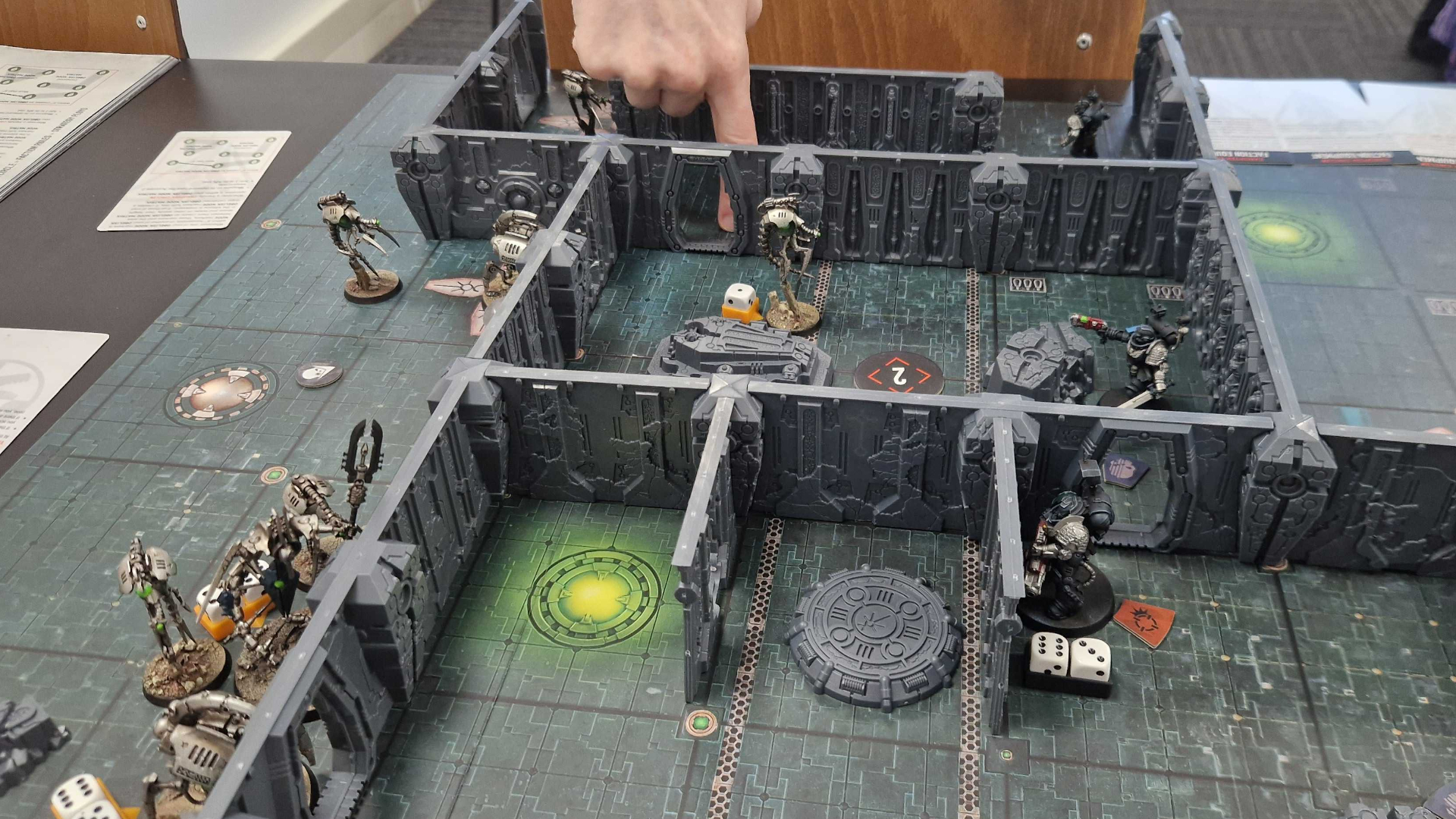A hand points to an in-progress board of Kill Team: Tomb World models, terrain, tokens, and cards