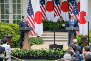 two men in suits stand at lecterns outdoors in front of four japanese and four american flags