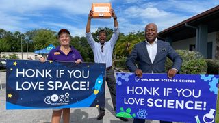 two people hold signs as a third person, in back, holds up a kit. the signs read "honk if you love space" and "honk if you love science" and have cartoon illustrations showing beakers and other science symbols