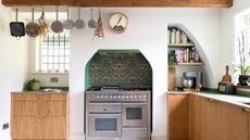 Kitchen cabinet trends are useful to know. Here is a kitchen with wooden cabinets, a silver oven with a green floral wall around it, a colorful bookshelf, and silver pots and pans hanging from the ceiling beam