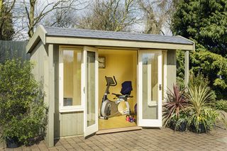 a small garden room with fitness equipment inside, with white doors and a green/grey exterior paint