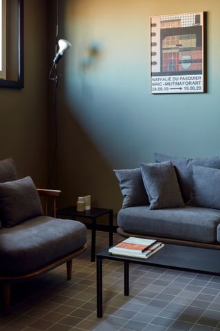 A grey living room with a grey couch and chair, a black coffee table and side table and dark grey floor tiles.