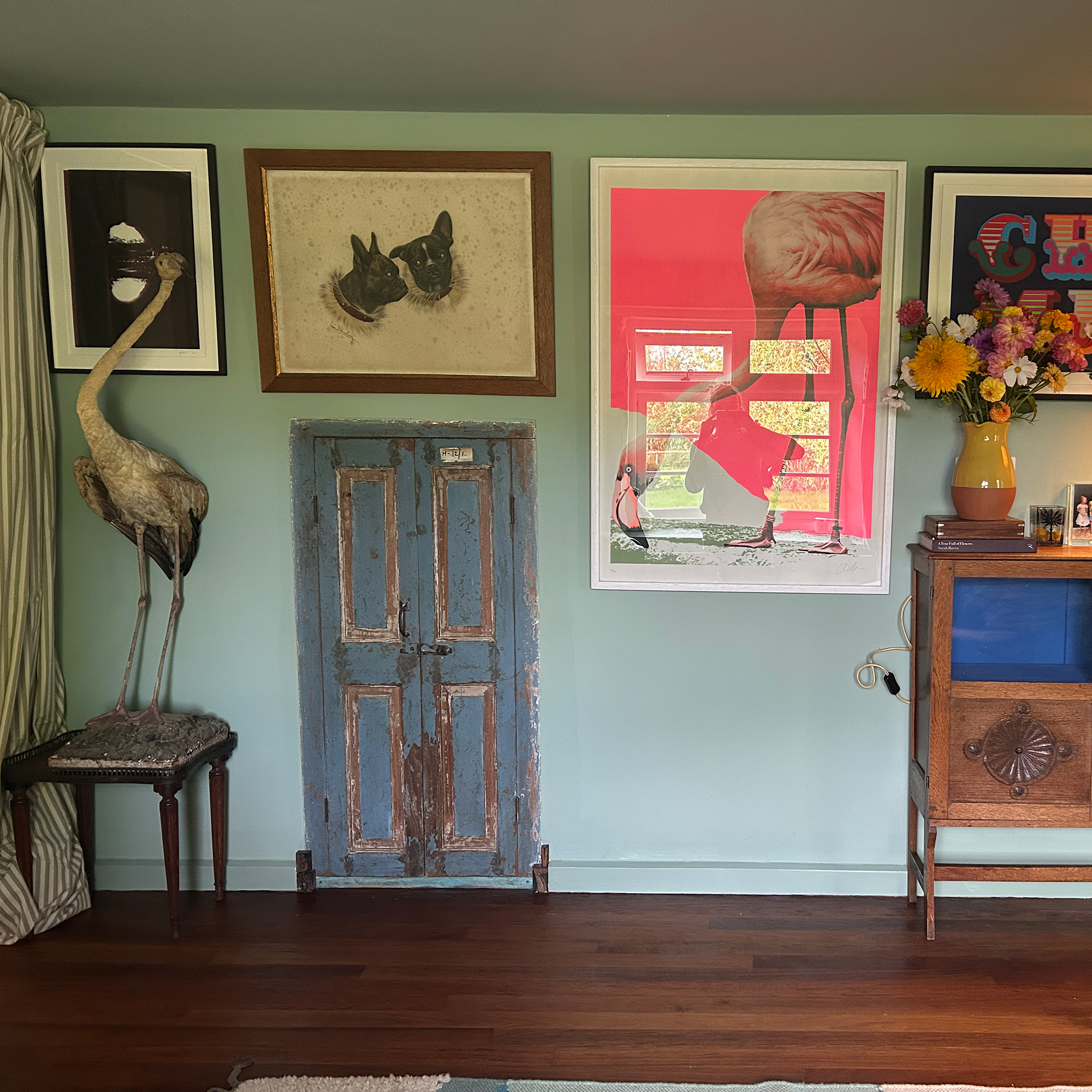 Green wall with small blue door, collection of large framed prints, heron on stool and wooden cabinet