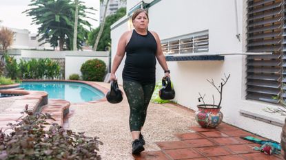 woman wearing workout clothes carrying a kettlebell in each hand walking outside with a swimming pool behind her. 