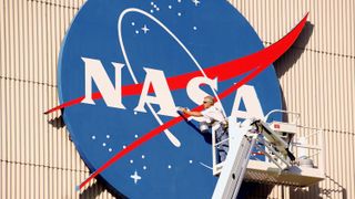 a person stands on a ladder and reaches towards a large nasa logo pasted on the outside of a building