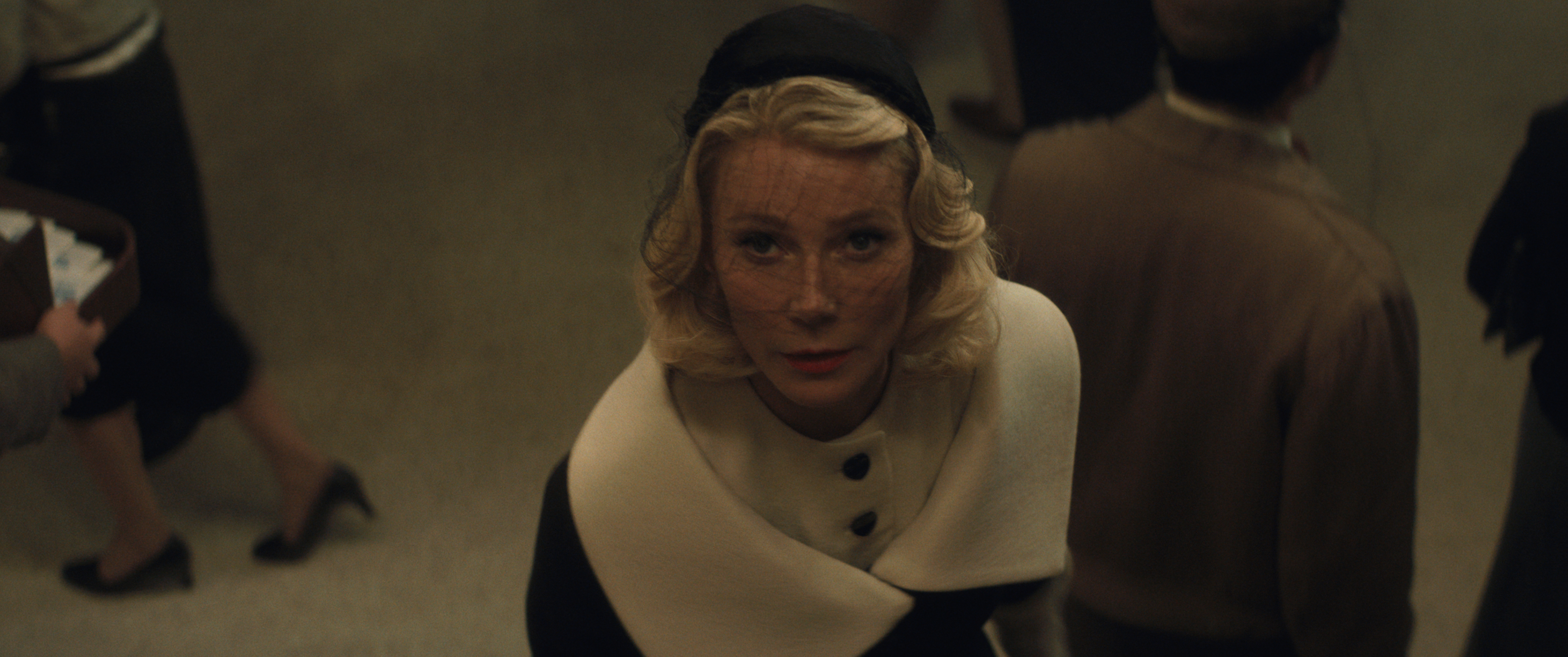 gwyneth paltrow as kay stone wearing a black and white regal coat and 50s era hat and veil looking upwards in marty supreme