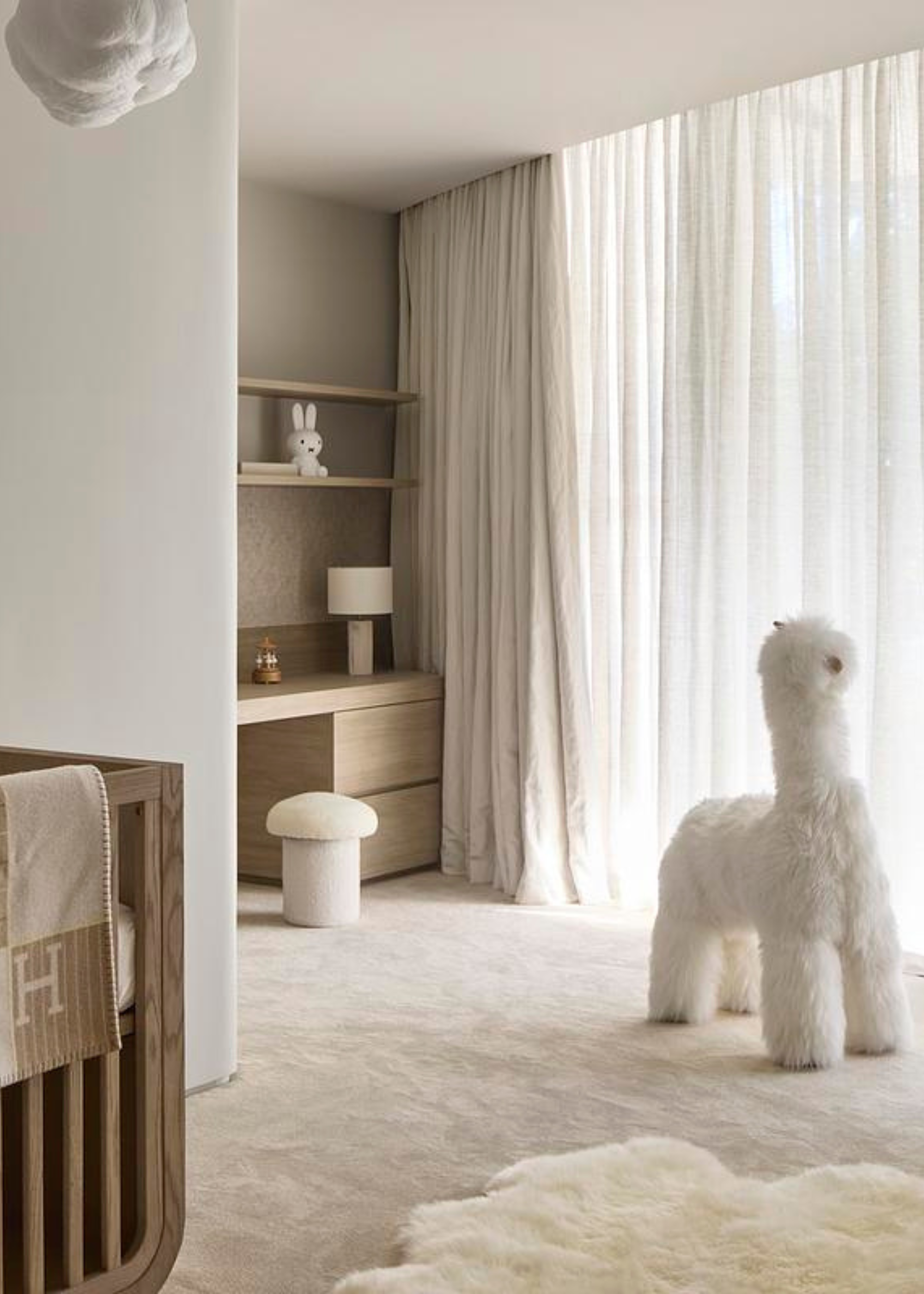 A minimalist kids room with a fuzzy alpaca doll, a mushroom stool by a wooden study with shelves, a wood crib with an Herm&amp;egrave;s blanket, and a fuzzy rug