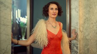 Nancy Mitford (Bessie Carter) stands in the doorway of a bar, with her hands touching either side of the door frame. She is wearing a low-cut red dress with a white feathery shawl draped around her shoulders.