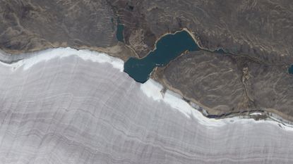 Satellite photo showing a lake bisecting the snowy edge of a gray ridged glacier