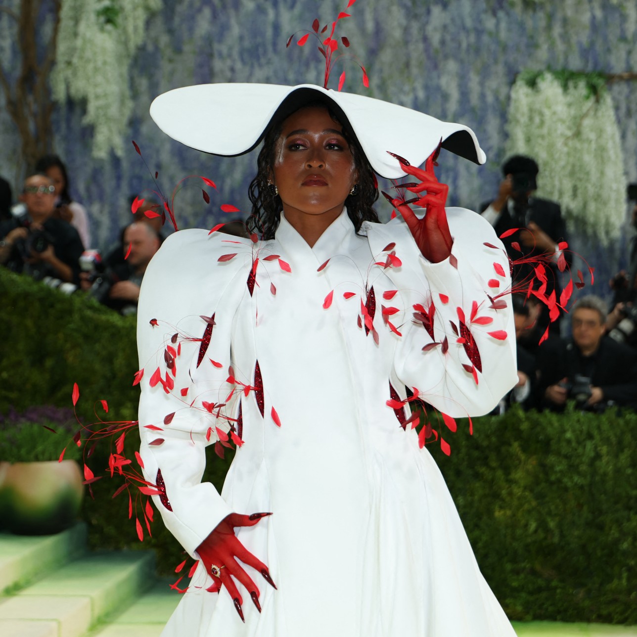 a woman attends the met gala in a long, white coat and a white hat with red feathers