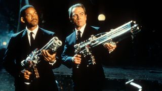 Screenshot from the movie Men in Black. Two men wearing black suits, white shirts, and a black tie are pointing rather large, futuristic looking guns to an off-screen target.