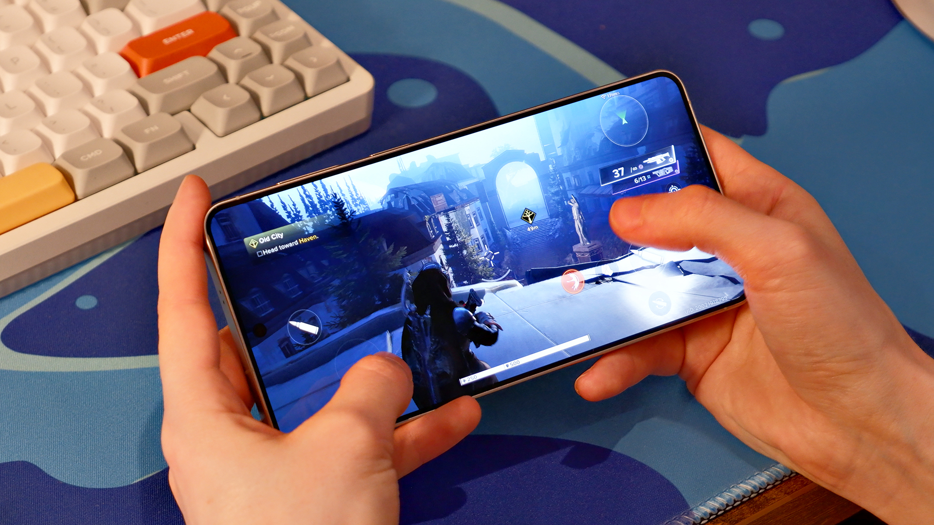 The Samsung Galaxy S26 Plus held in hand, playing Destiny Rising against a blue mousepad and white keyboard.