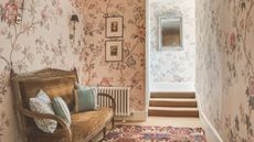 A pink floral wallpapered hallway with a vintage upholstered bench alone one wall. 