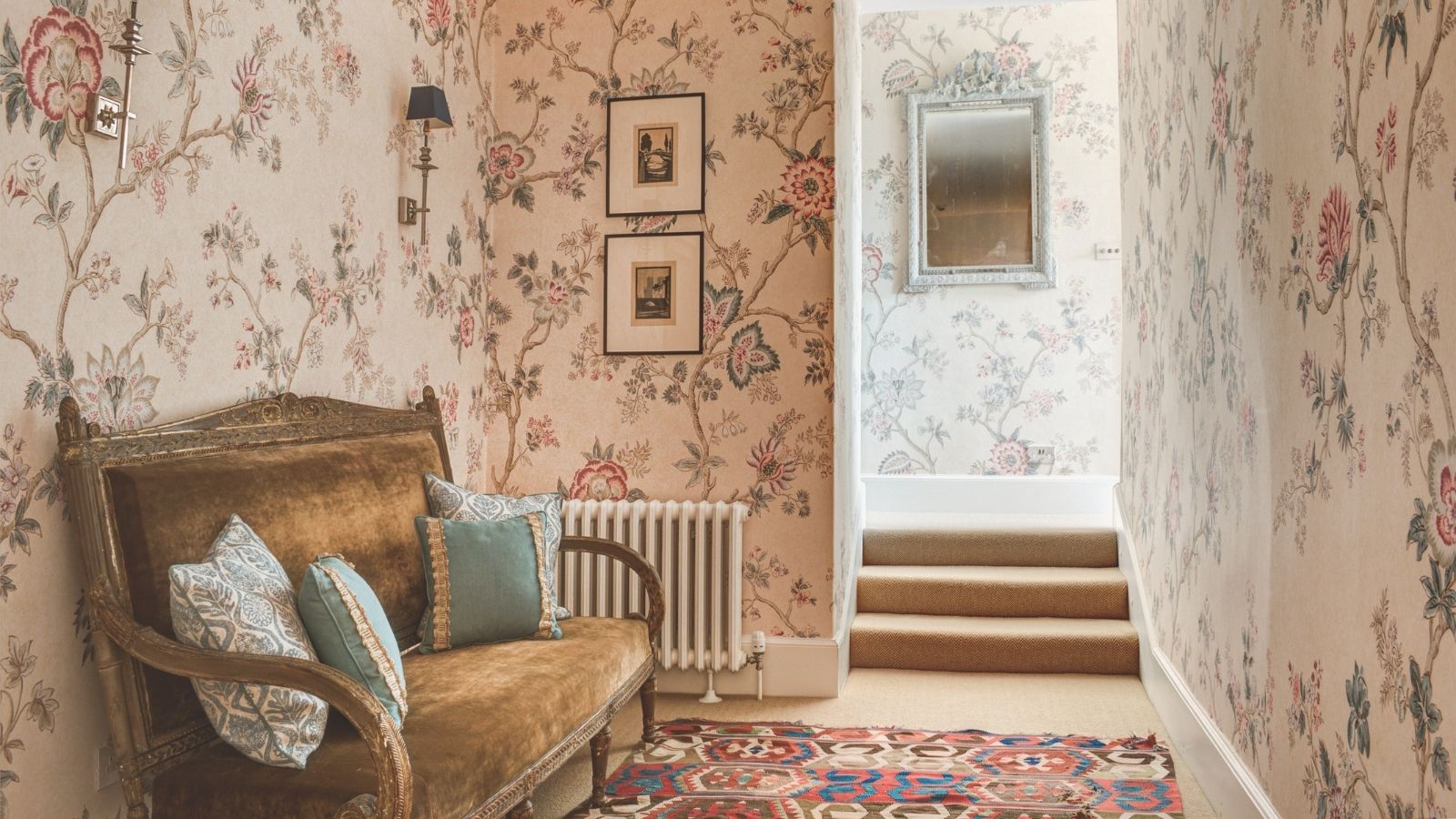 A pink floral wallpapered hallway with a vintage upholstered bench alone one wall. 