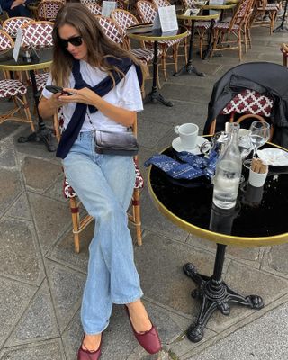 A French woman sitting outside at a café wearing jeans, a white tee, burgundy ballet flats, and black sweater around her shoulders