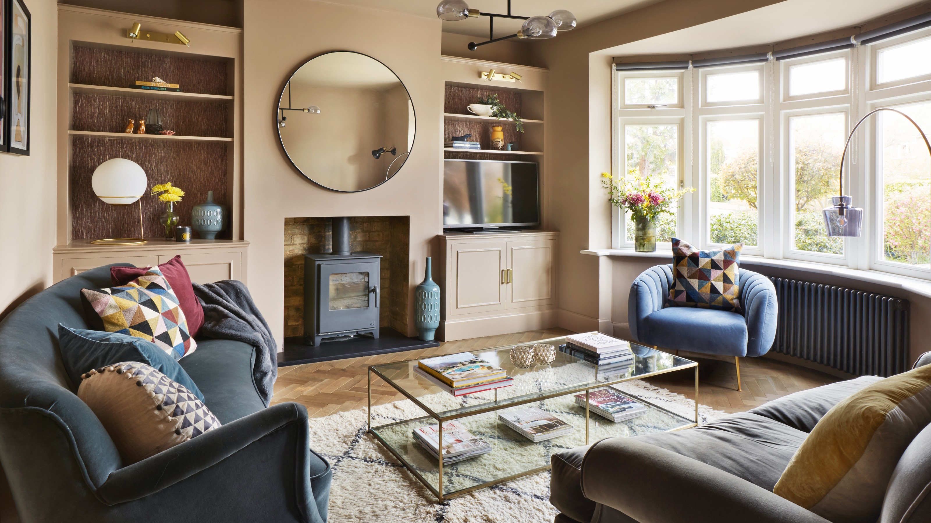 Cosy warm neutral living room with bay window, alcove storage, log burner, circle mirror, glass coffee table, textured rug and accent chair next to radiator