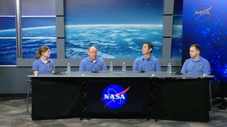 Found individuals wearing pale blue shirts sit behind a long table with a black cloth over it and the blue NASA circle logo with blue screens behind them.