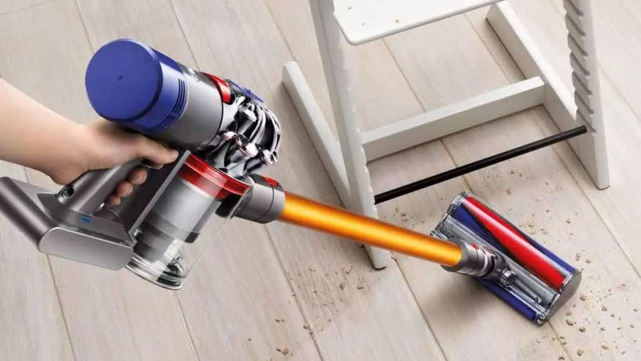 Hand holding Dyson V8 and vacuuming cereal and small debris from a hardwood floor next to a chair.