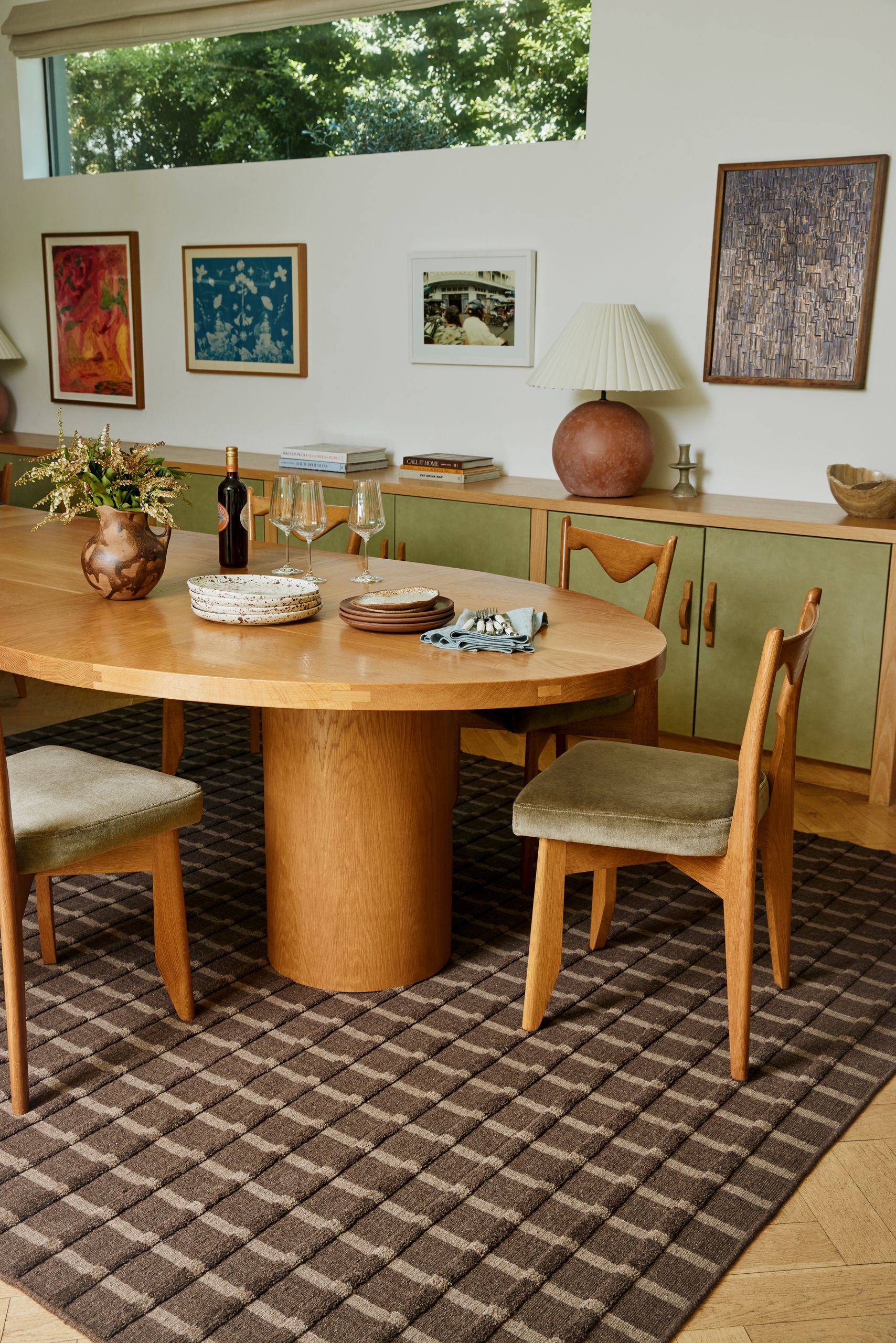 A brown wool rug under a large, oval dining table with wooden dining chairs with green velvet seats. There is a long green and wooden console across the back wall. 
