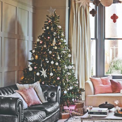 A living room with wall panelling decorated for Christmas with a large Christmas tree in the corner fully decorated with lights, ornaments and a tree topper