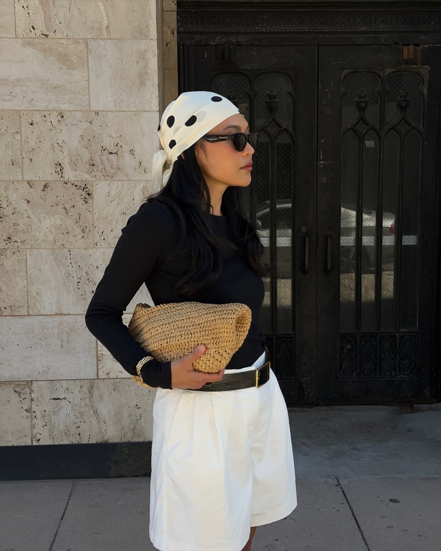 Woman wears polka dot scarf, black top, white shorts and raffia bag