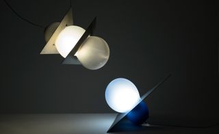 Two large vase bulbs pointing towards each other. One is resting on a table and the other is hanging down. On the table one has a blue illumination. The one above illuminates in a beige colour.