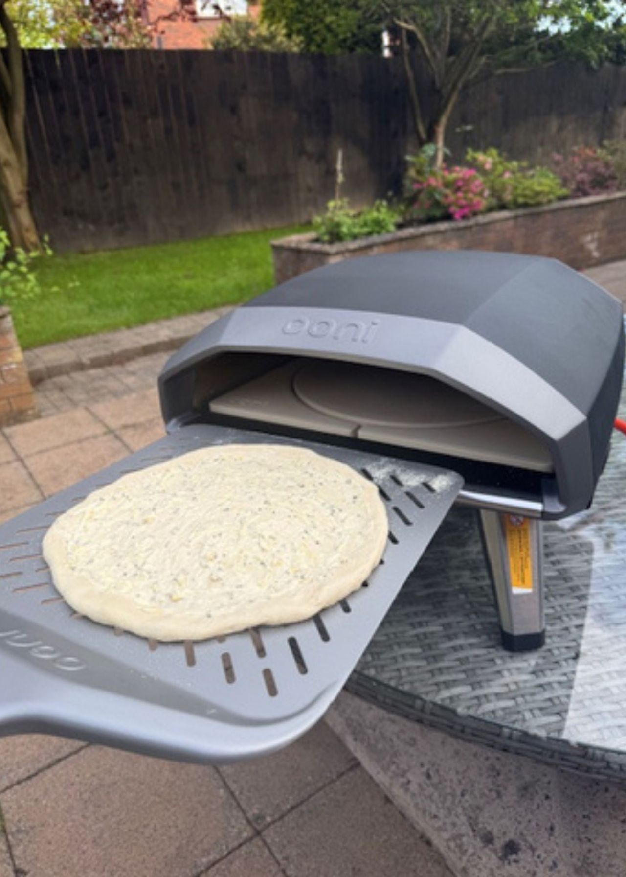 A peel with a prepared dough is being put into an Ooni Koda 2 pizza oven with a rotating stone on a patio with flower beds and grass in the background