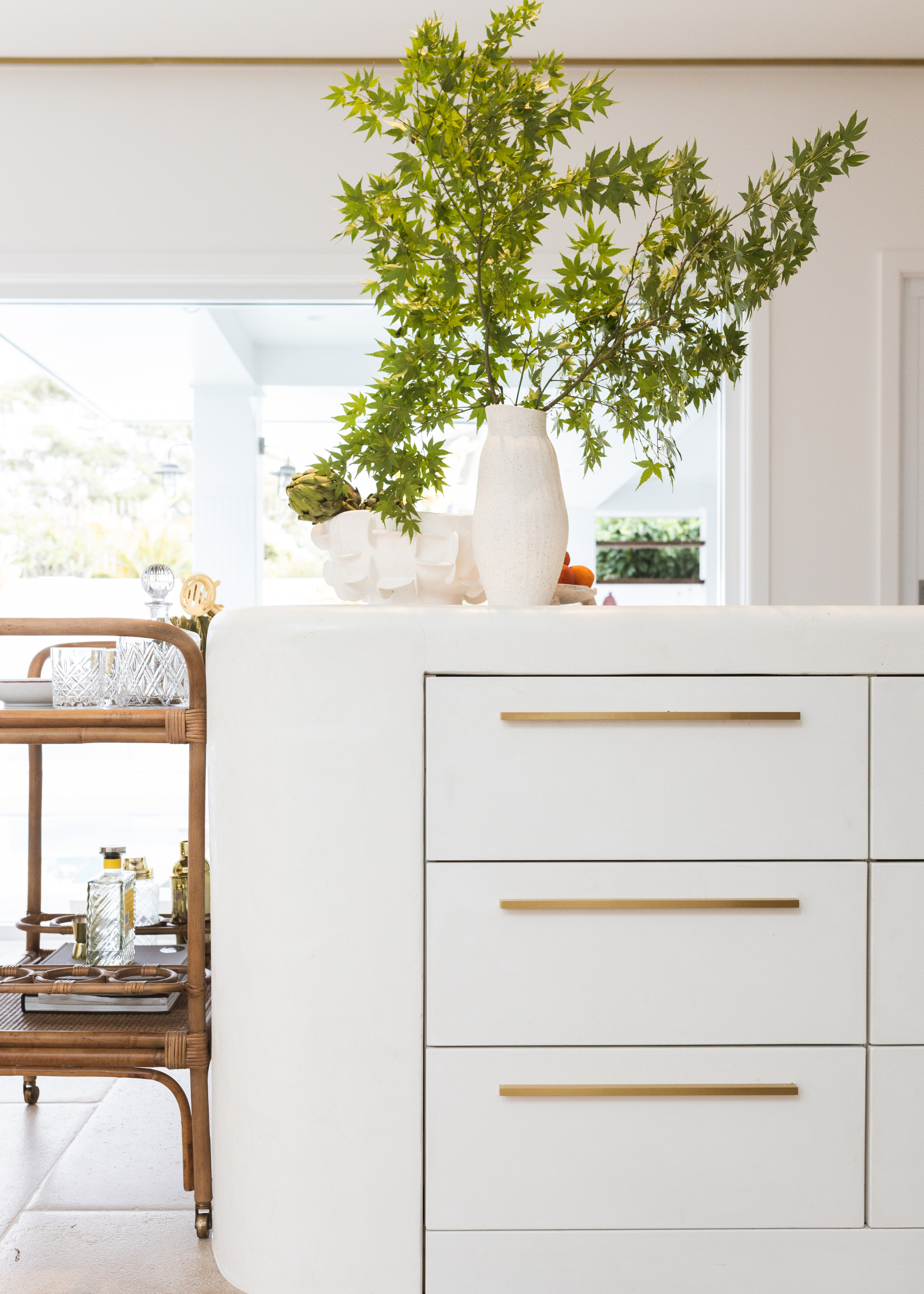A white kitchen island with drawers featuring gold hardware. A vase with stems on the counter beside a bamboo-style trolley with drinkware
